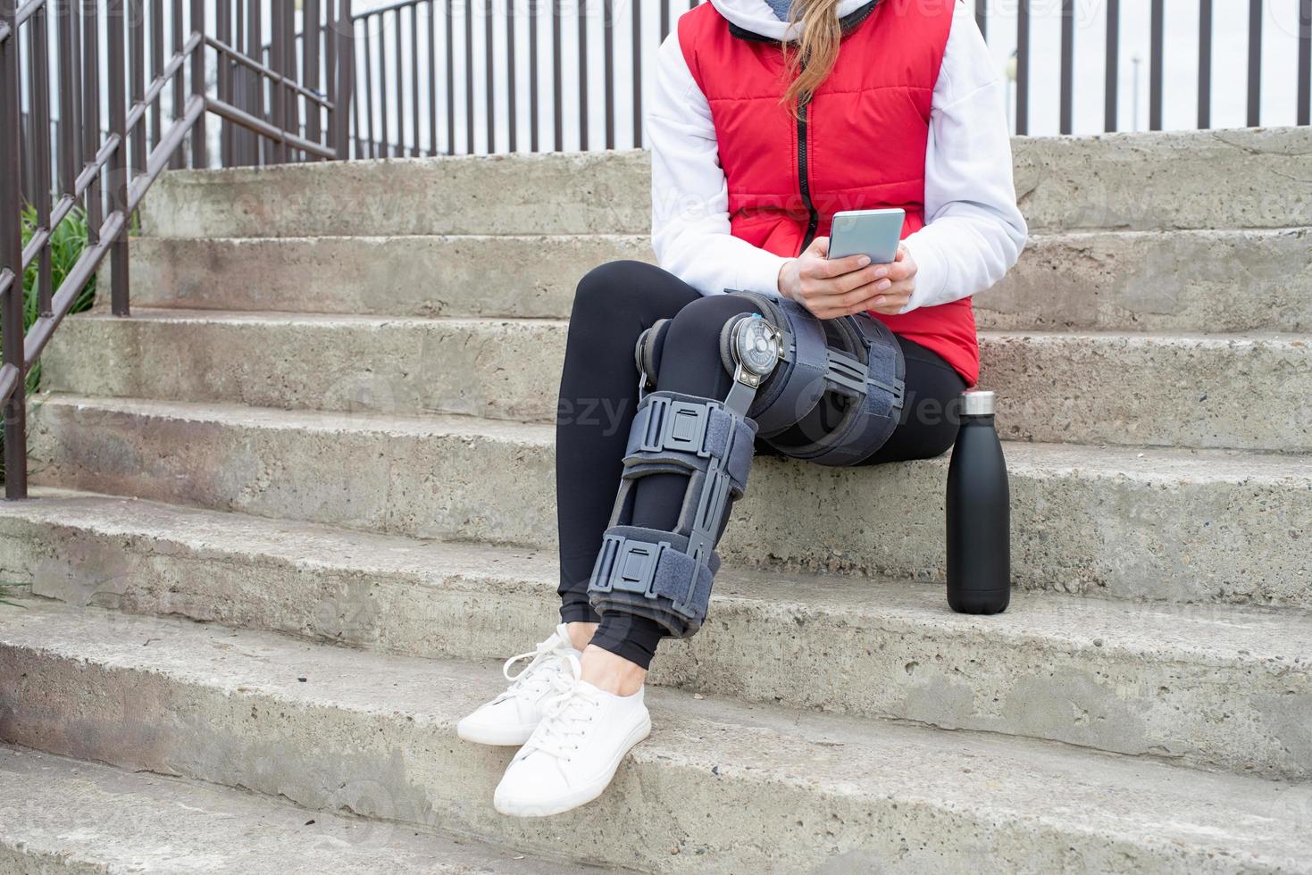 Woman wearing knee brace or orthosis after leg surgery, sitting on stairs ourdoors photo