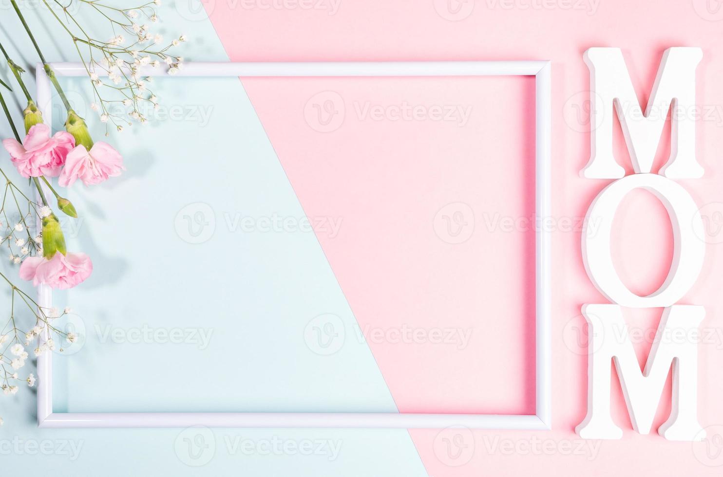 Empty white frame, pink carnations and gypsophila flowers, white letters MOM on pink- blue backdrop. photo