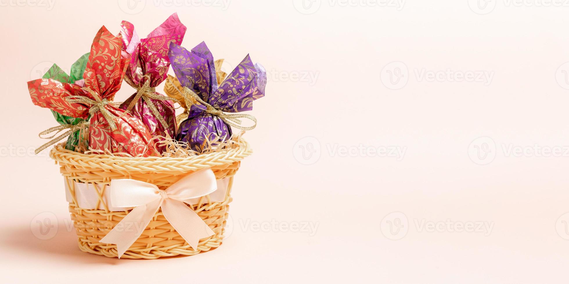 Easter eggs wrapped in color paper in basket decorated by bow on pastel pink backdrop. Banner. photo