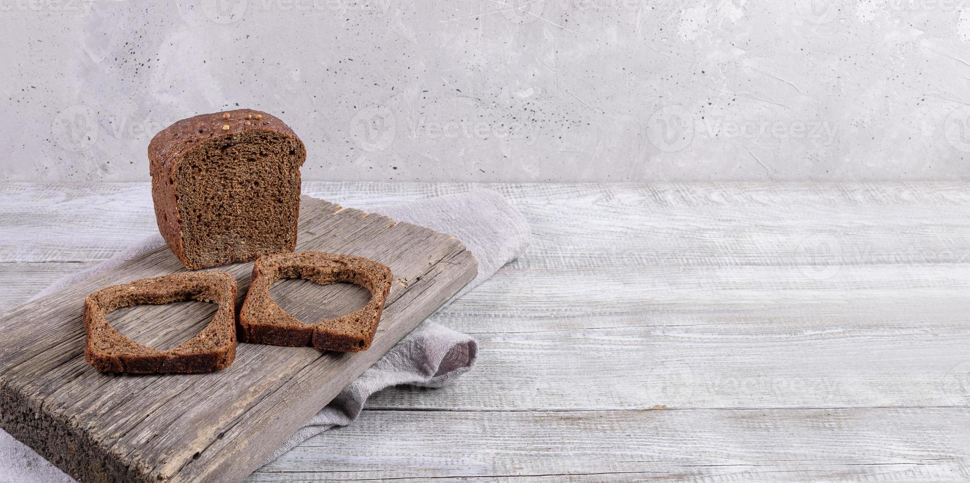 Loaf of rye bread and two slices with carved holes of heart shape in them on old wooden Board on gray background. photo