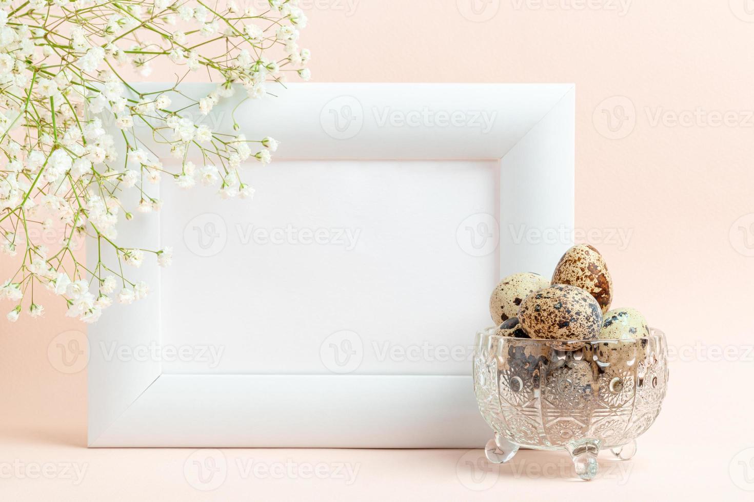 Empty white frame, quail eggs in crystal bowl and white gypsophila on pastel pink. Mock up. photo