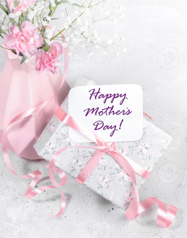 Wrapped gift box with ribbon, Happy mother's day note, pink vase with flowers on grey close up. photo