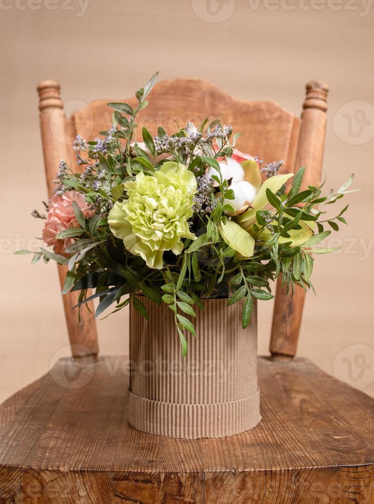 Eco friendly composition with flowers bouquet in DIY cardboard vase standing on old wooden chair on beige background. photo