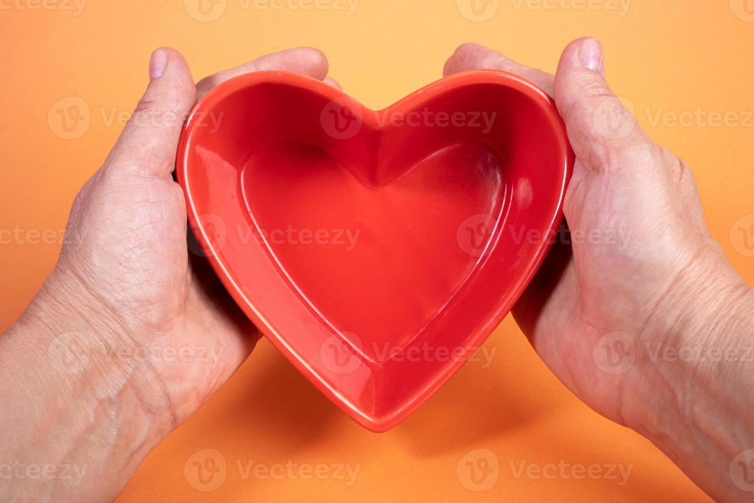 Woman hands holding small empty red plate in heart shape in orange background. Creative mockup. photo