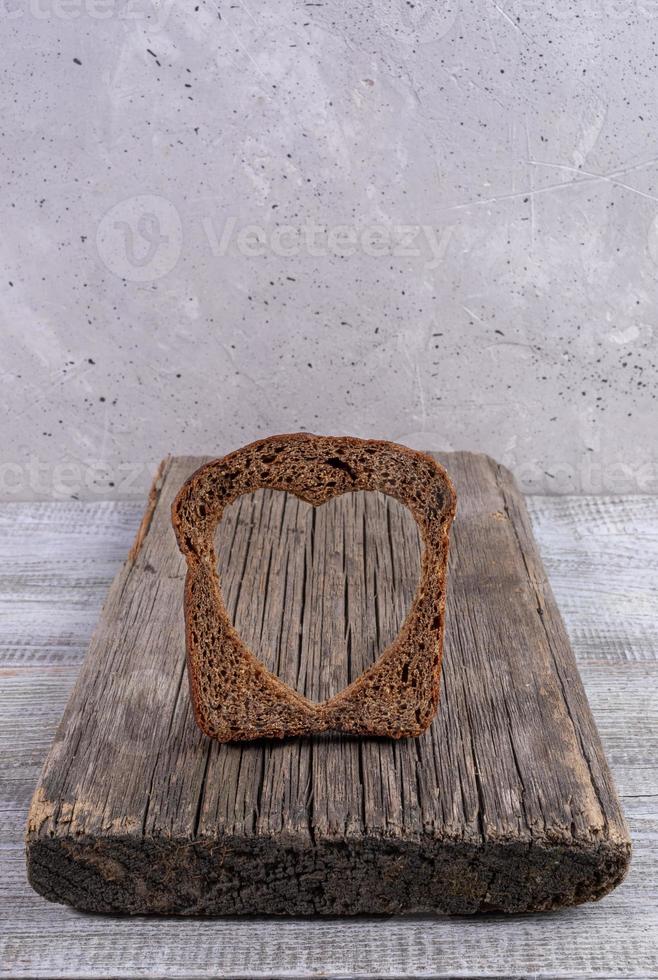 One rye bread slice with carved hole of heart shape in it on old wooden board on grey concrete backdrop. photo