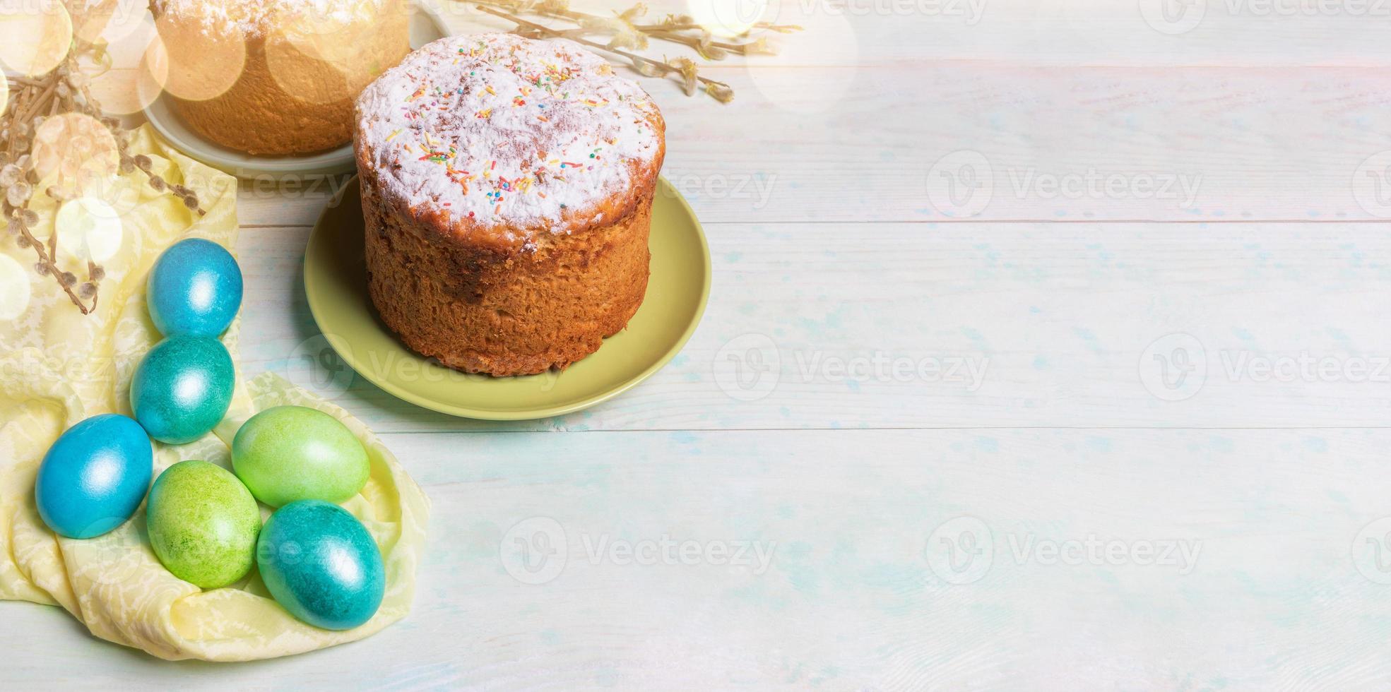 Easter festive banner with Easter cakes, colored Easter eggs on yellow cloth, willow twigs, bokeh on wooden background. photo
