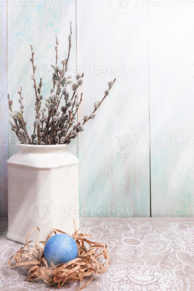 Easter blue egg in nest and willow branches in vase on wooden wall background with sunbeams overlay and copy space. photo
