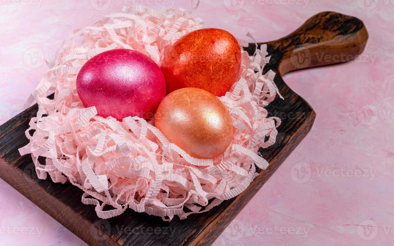 Three colored eggs in nest from pink paper shreds on dark wooden kitchen board on pink textured background. Close-up. photo