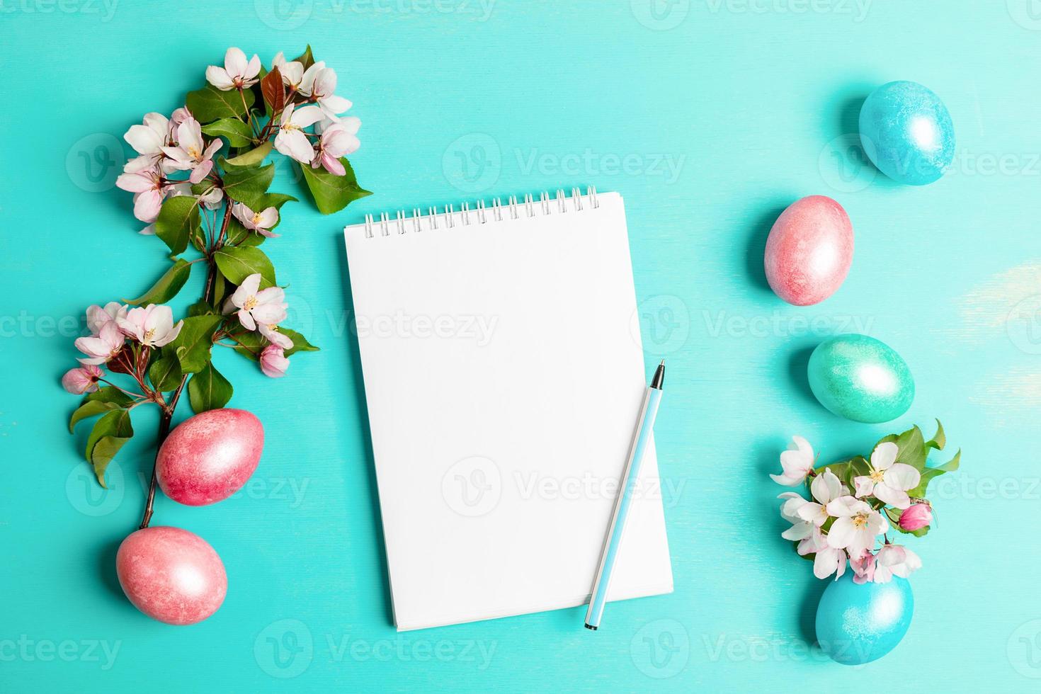 Festive Easter layout with pink Apple tree twigs, colored eggs, white blank notepad with pen on turquoise backdrop. photo