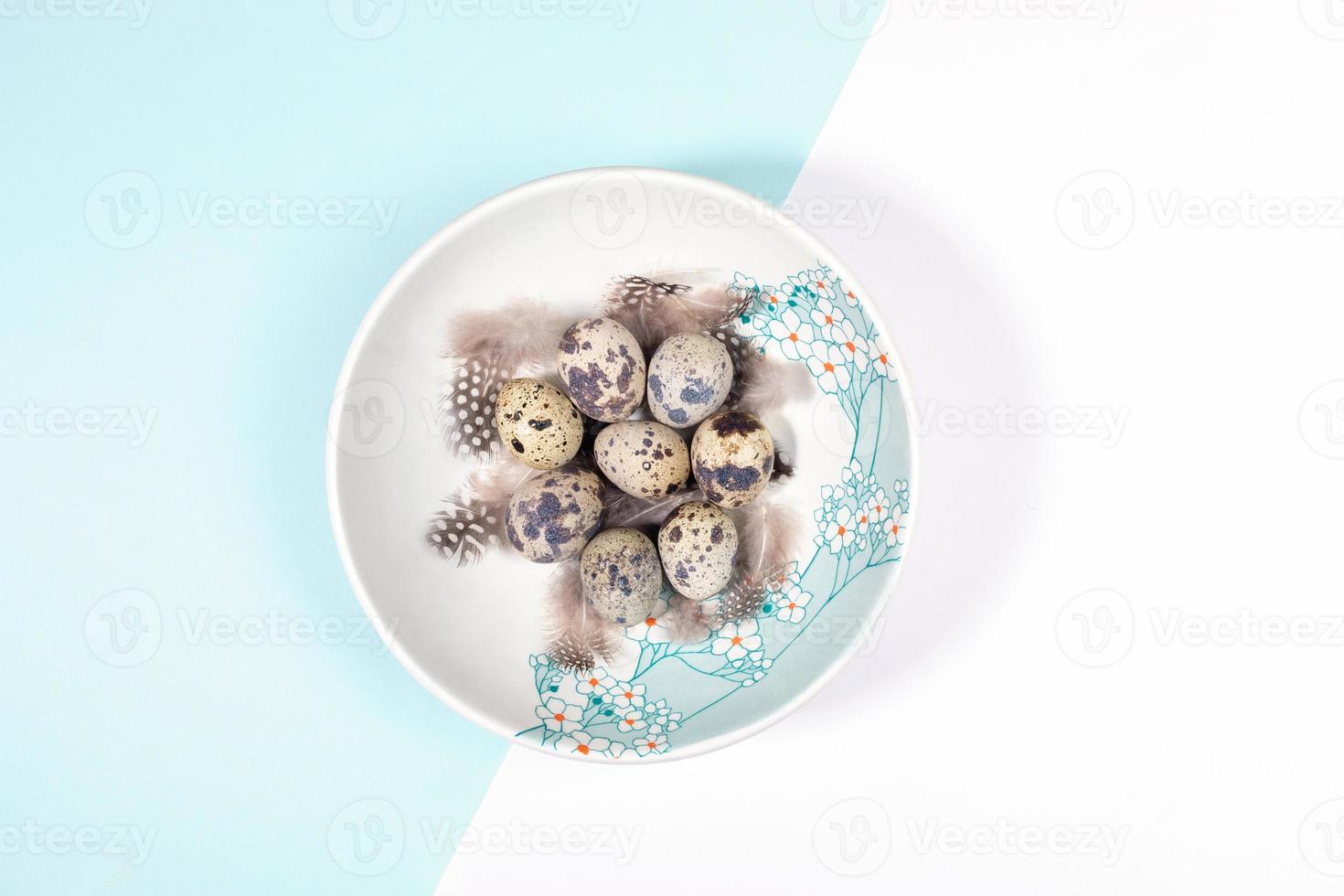 concepto de ajuste de la mesa de pascua con huevos de codorniz y plumas en un plato decorado con flores en blanco - fondo azul claro. foto