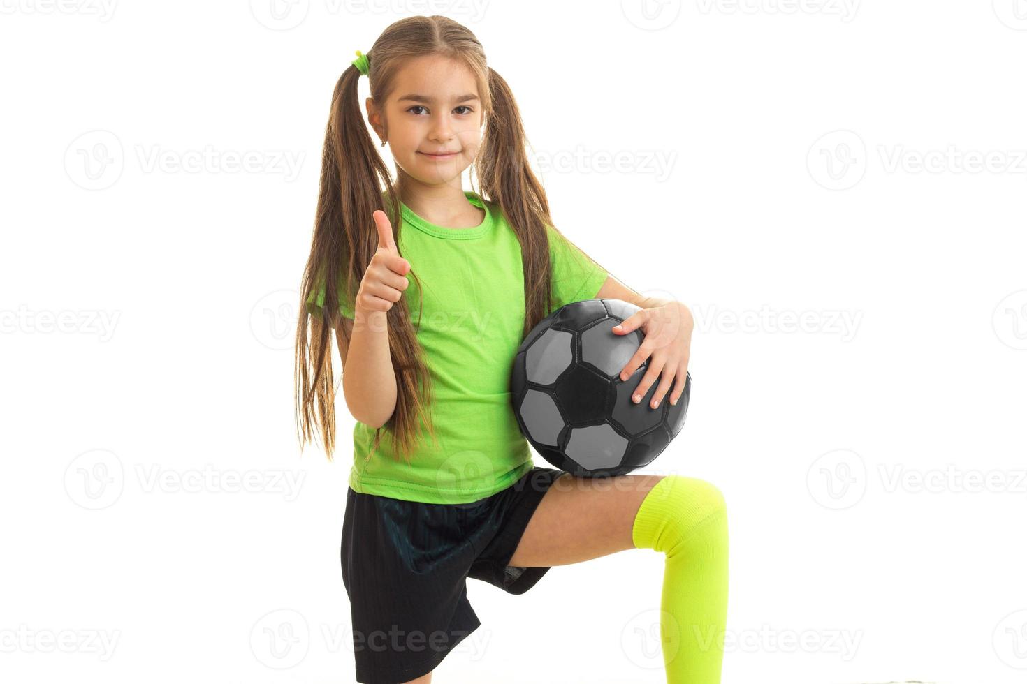 little girl in sports uniform with soccer ball in hands shows thumbs up photo