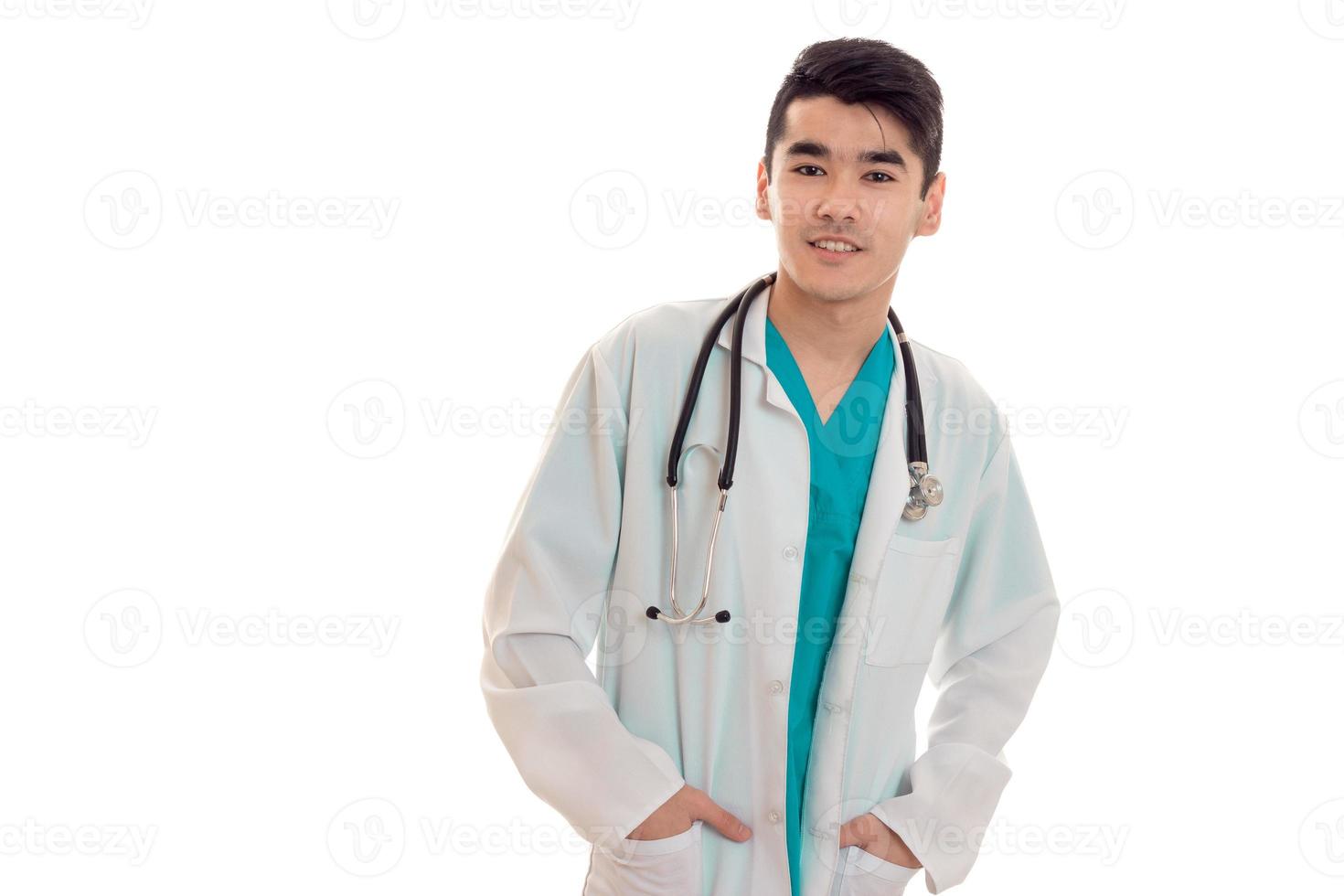 un joven médico con una bata blanca de laboratorio mantiene las manos en los bolsillos y sonríe aislado de fondo blanco foto