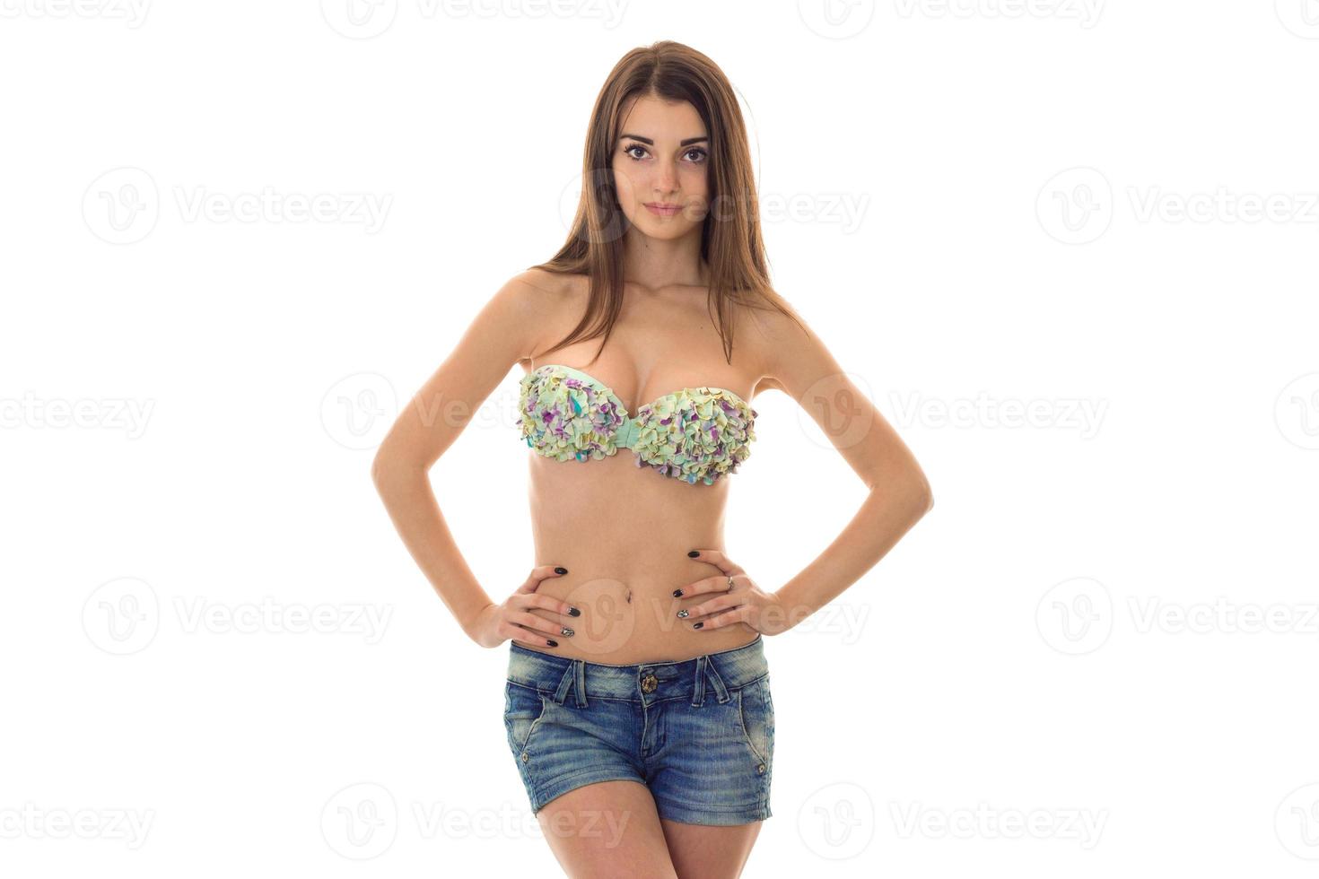 young cheerful brunette woman with big natural breasts in swimsuit with floral pattern and jeans shorts looking at the camera with hands on her hips isolated on white background photo