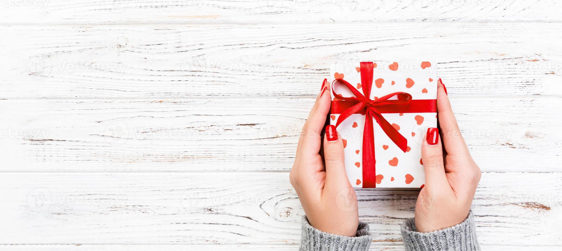 Woman hands give wrapped valentine or other holiday handmade present in paper with red ribbon. Present box, red heart decoration of gift on wooden table, top view with copy space for you design photo