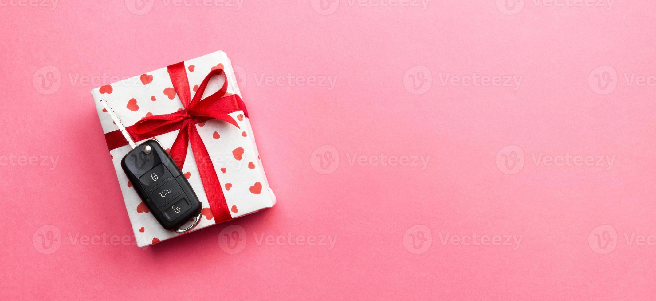 Car key on paper gift box with red ribbon bow and heart on pink table background. Holidays present top view concept photo