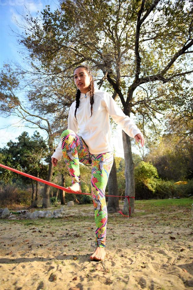 Woman balancing her walk on a loose rope tied between two trees. Woman practicing slack rope walking in a park. photo
