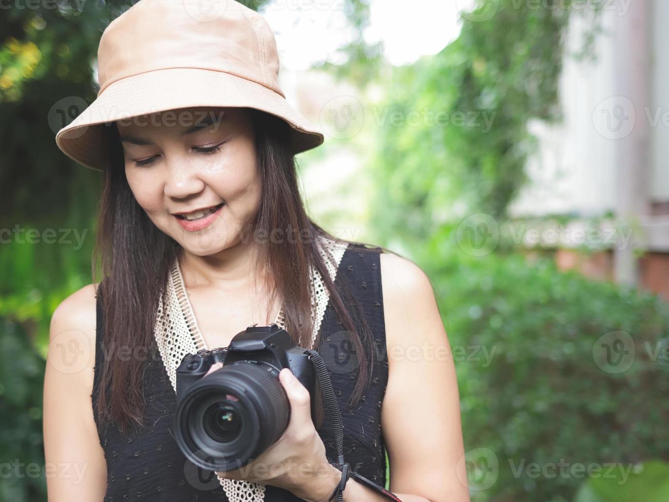 mujer asiática, con sombrero y blusa negra sin mangas, parada en el jardín y sosteniendo una cámara dslr, sonriendo alegremente y mirando hacia abajo. foto