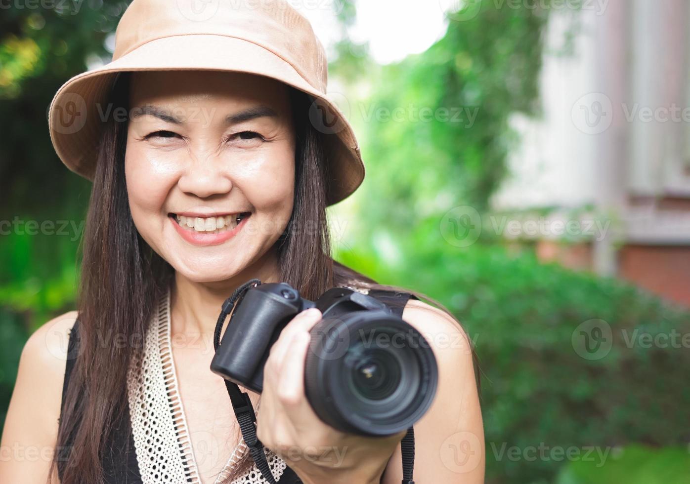 mujer asiática, con sombrero y blusa negra sin mangas, parada en el jardín, sosteniendo una cámara dslr, sonriendo alegremente. foto