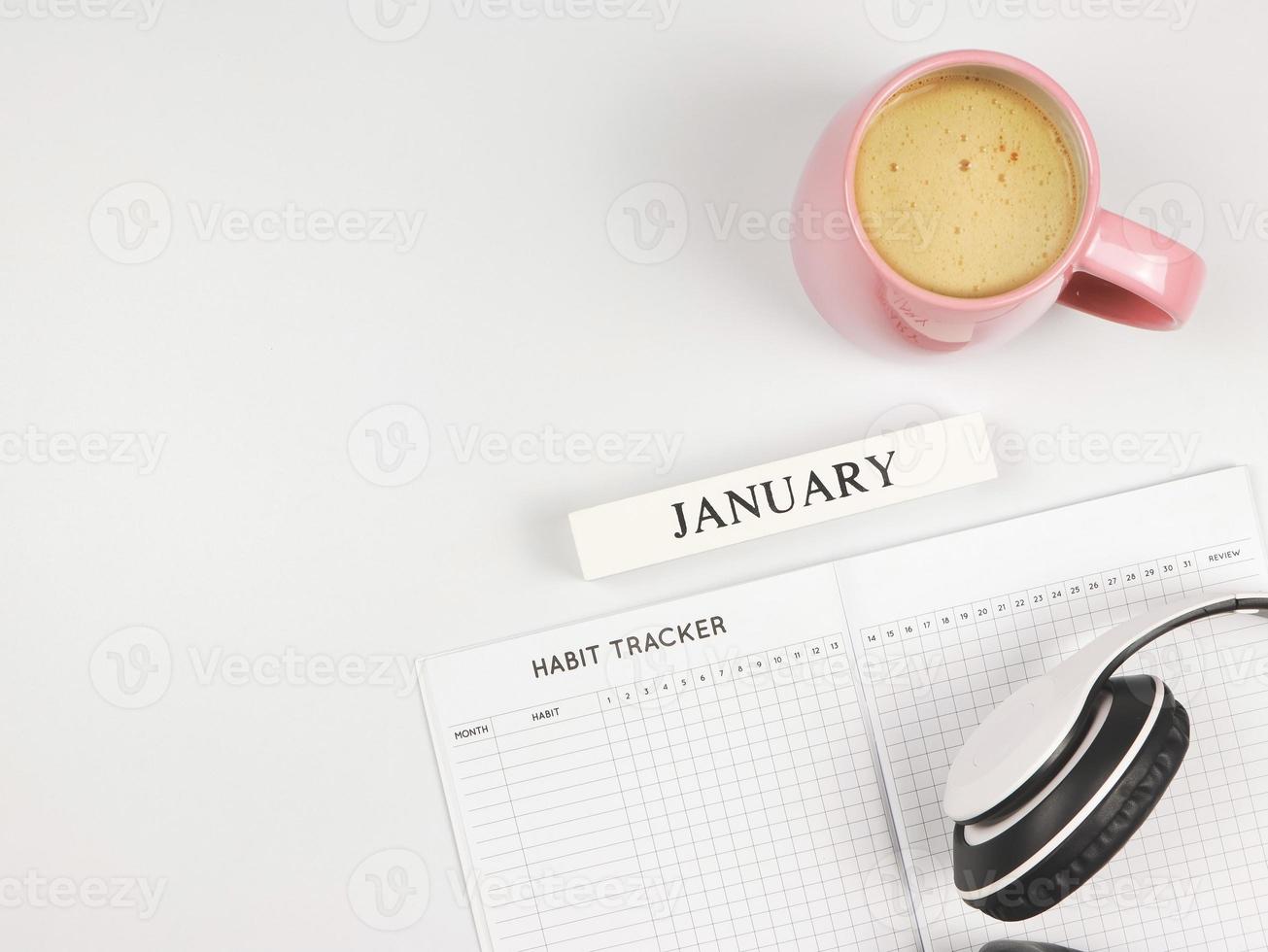 flat lay of habit tracker book, wooden calendar January, pink cup of coffee and headphones on blue background with copy space. photo