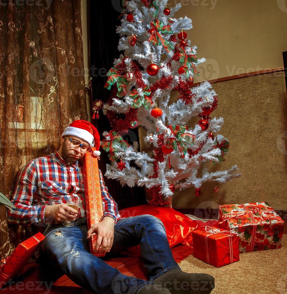 Drunk nerd in santa hat lying under the Christma tree with a lot of presents photo