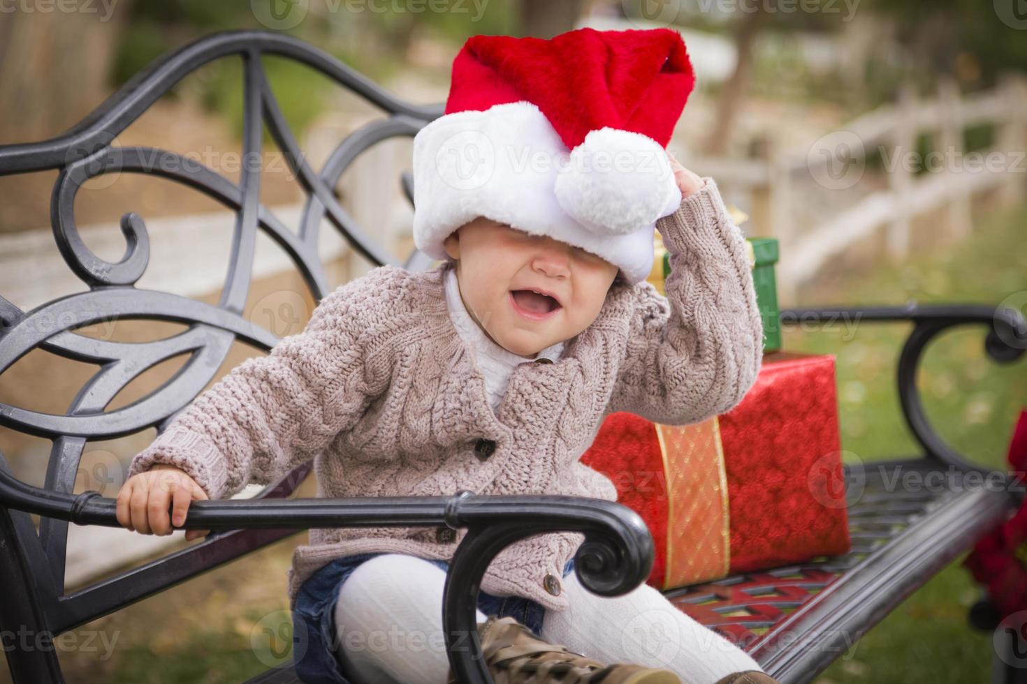 Young Child Wearing Santa Hat Sitting with Christmas Gifts Outside. photo