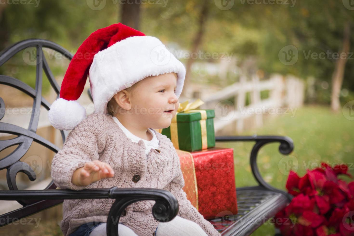 Young Child Wearing Santa Hat Sitting with Christmas Gifts Outside. photo