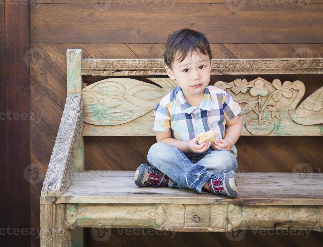 Cute Mixed Race Boy Sitting on Bench Eating Sandwich photo