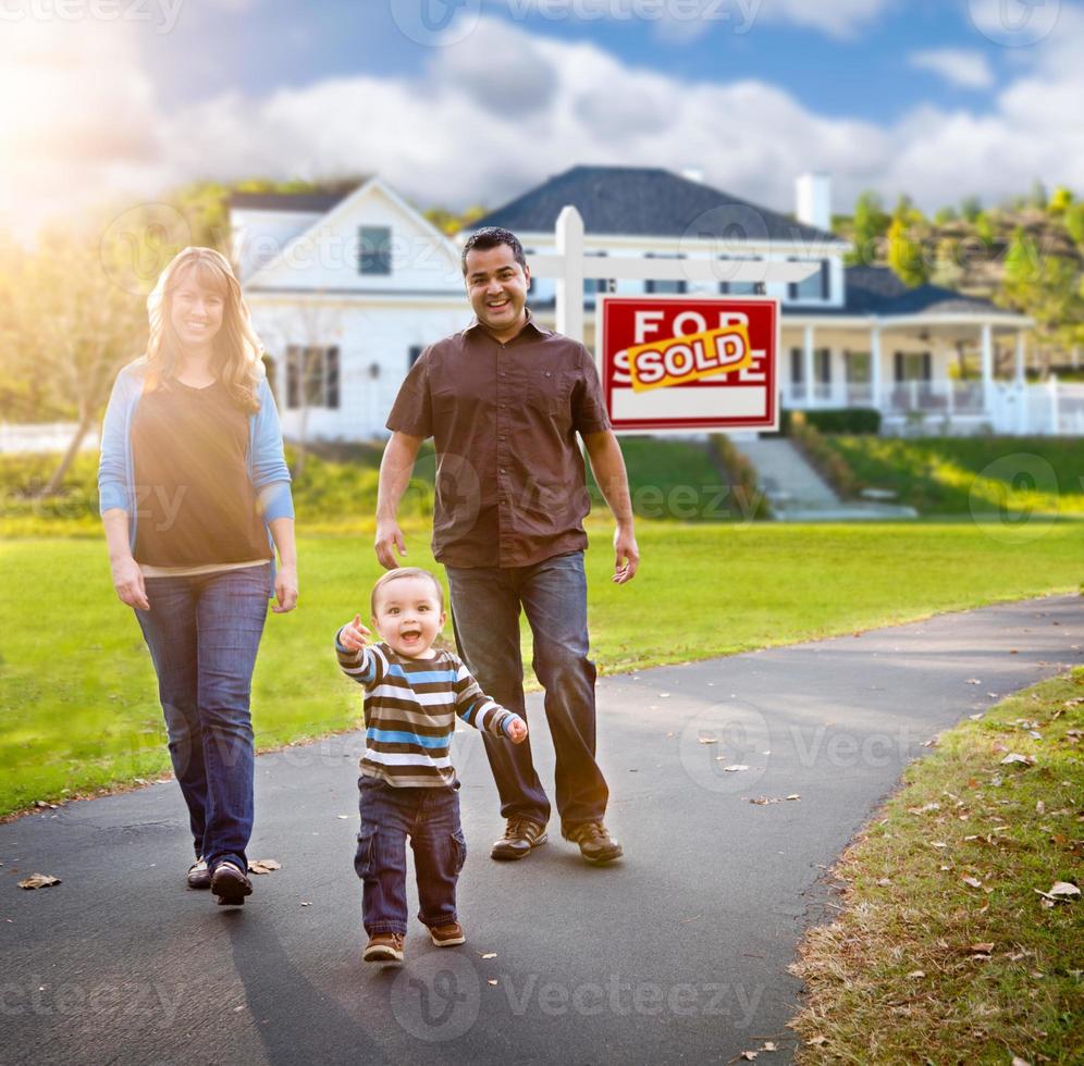 feliz familia de raza mixta caminando frente a casa y vendido en venta letrero de bienes raíces. foto