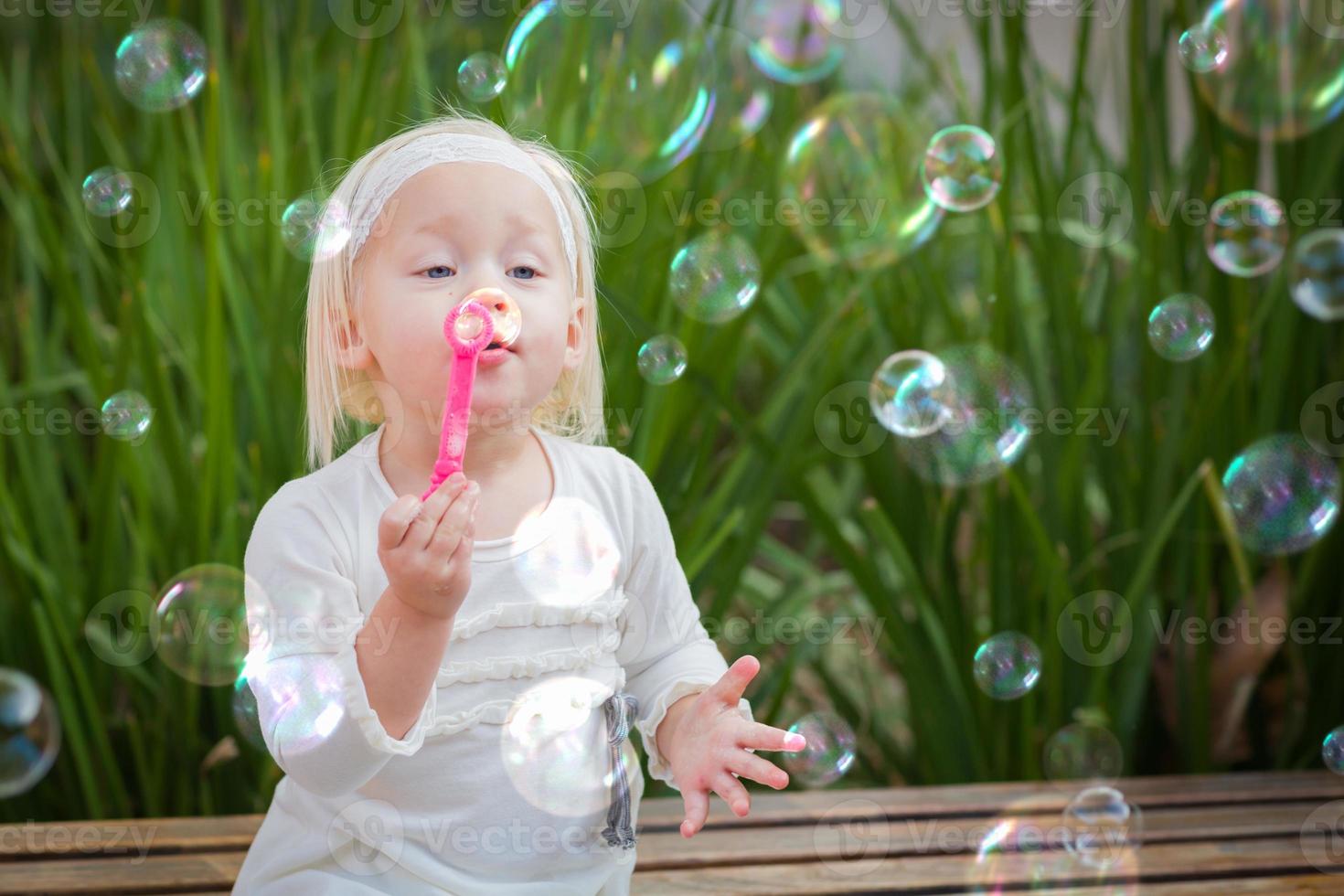 Adorable Little Girl Sitting On Bench Having Fun With Blowing Bubbles Outside. photo