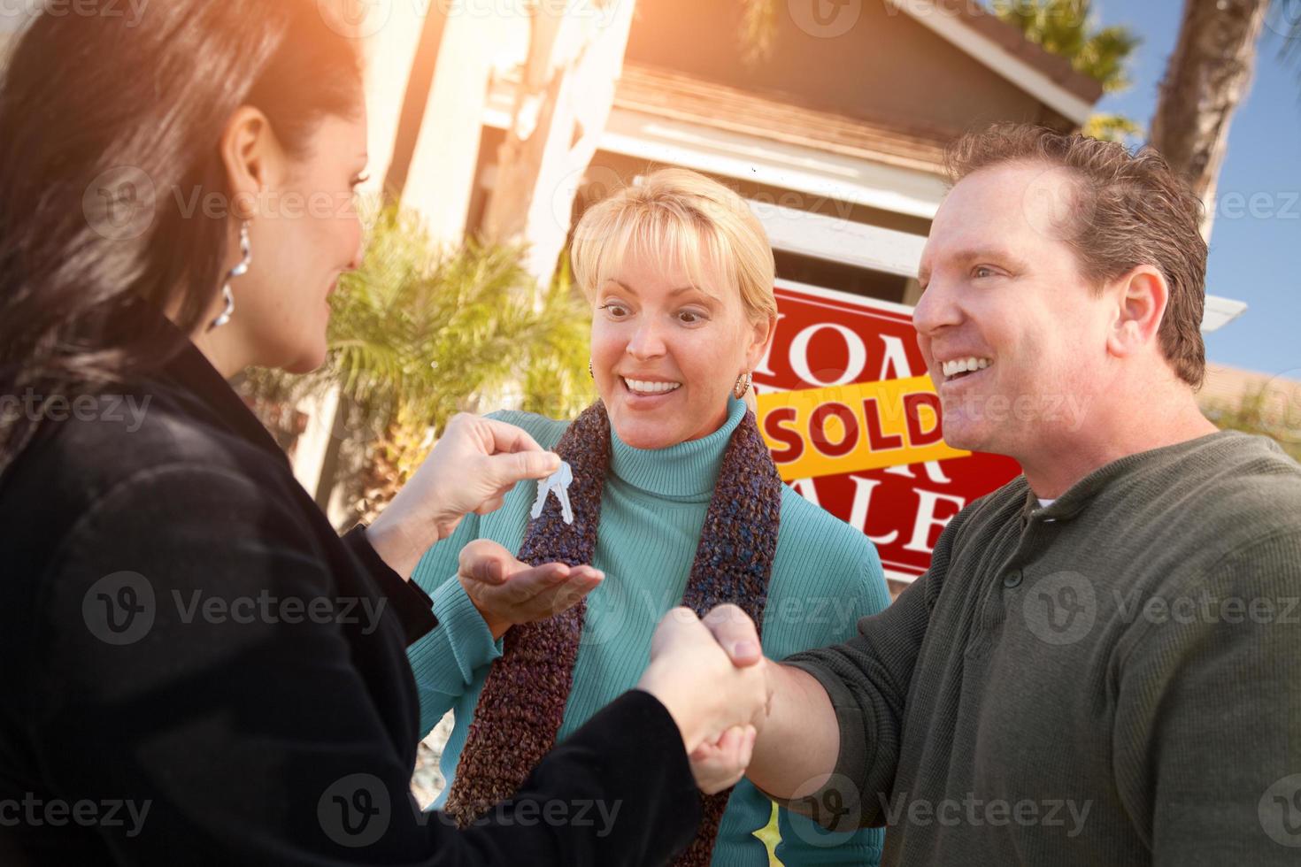 Hispanic Female Real Estate Agent Handing Over New House Keys to Happy Couple In Front of Sold For Sale Real Estate Sign photo