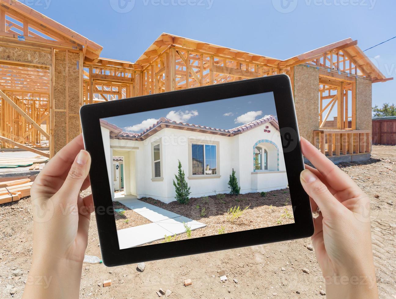 Female Hands Holding Computer Tablet with Finished House on Screen, Construction Framing Behind. photo
