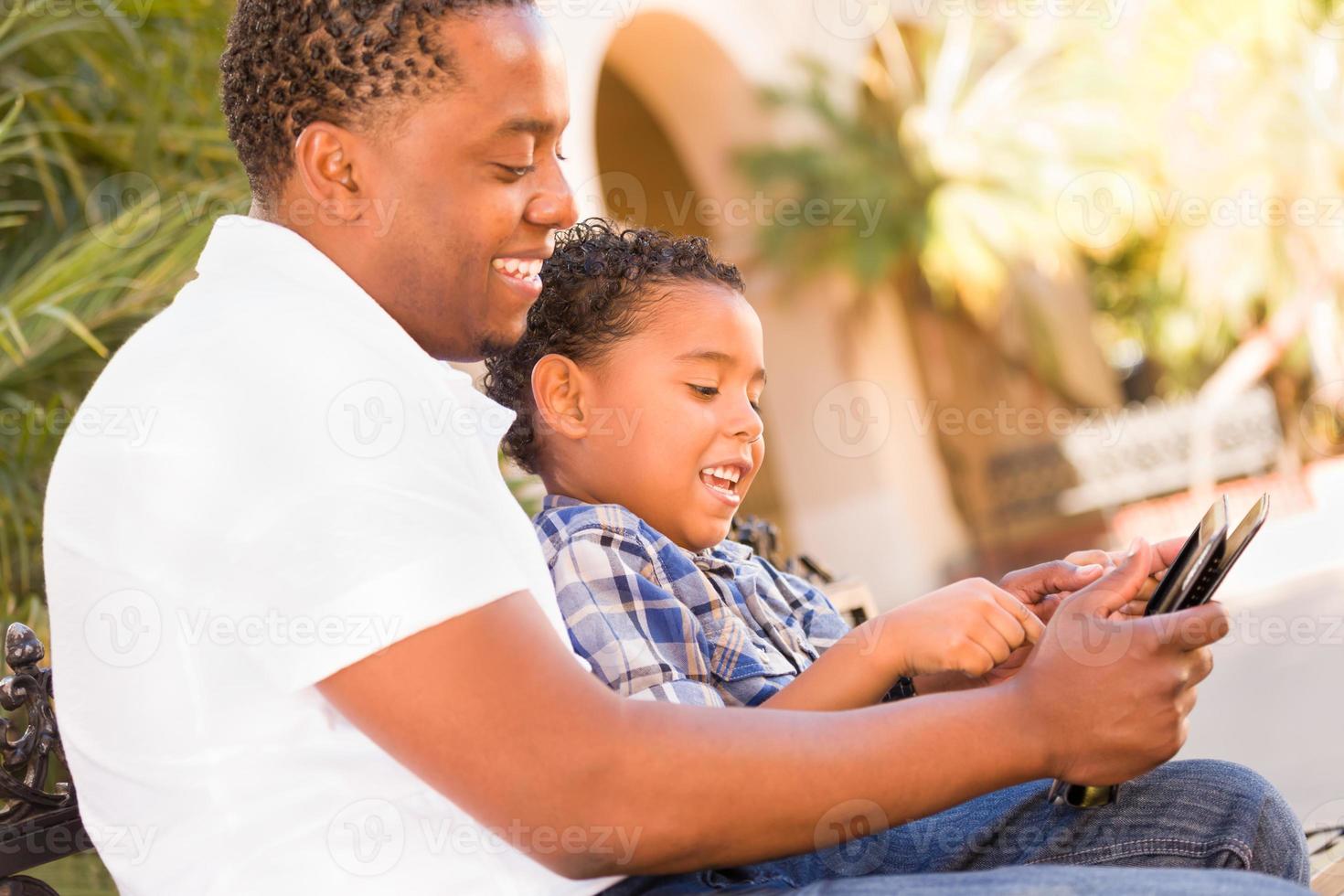 African American Father and Mixed Race Son Using Computer Tablet on Bench in Park photo