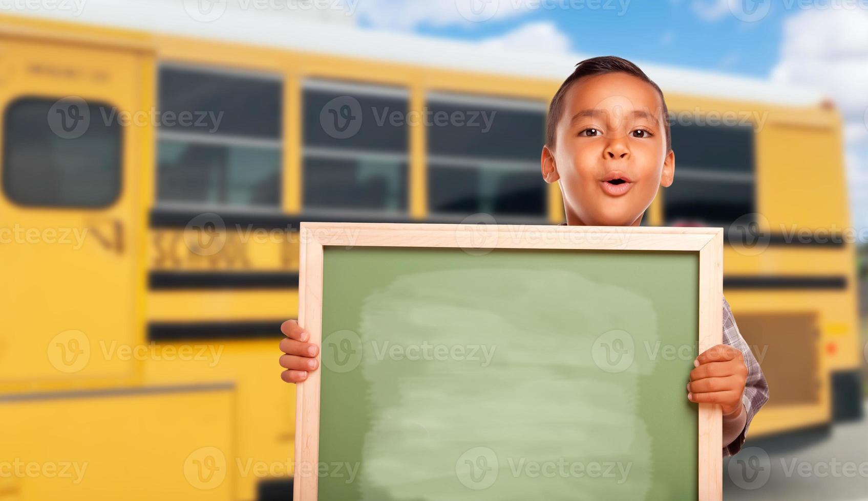 Young Hispanic Boy with Blank Chalkboard Near School Bus photo