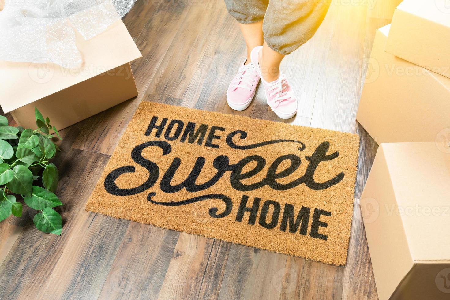 Woman in Pink Shoes and Sweats Standing Near Home Sweet Home Welcome Mat, Boxes and Plant photo