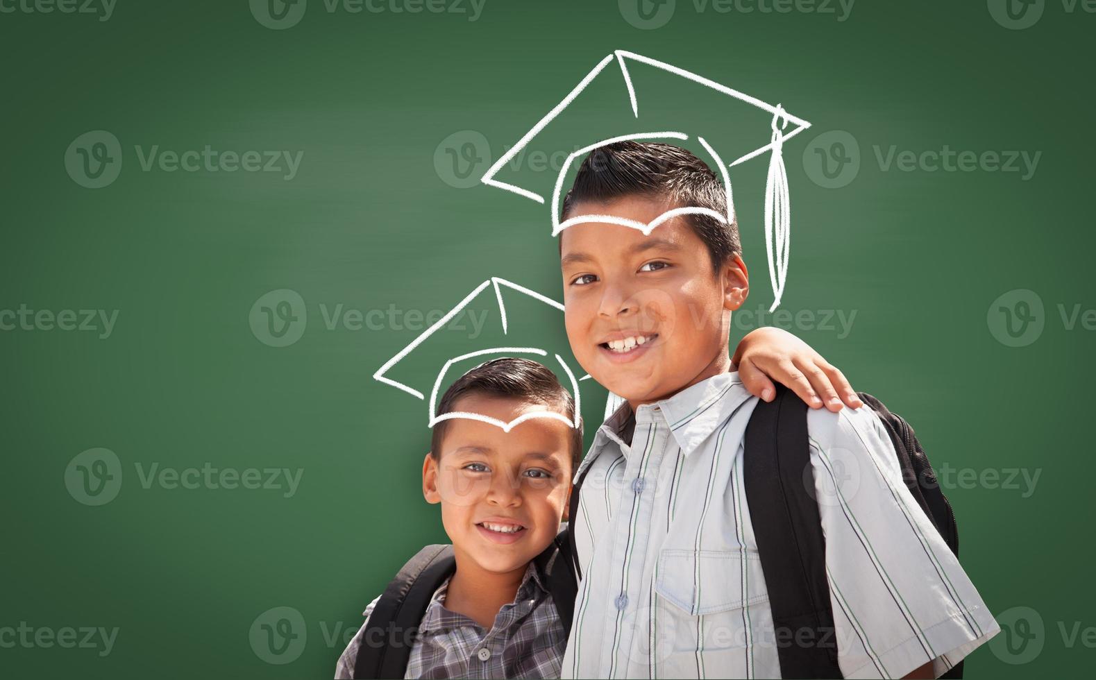 Young Hispanic Student Boys Wearing Backpacks In Front Of Blackboard with Graduation Caps Drawn In Chalk Over Head photo