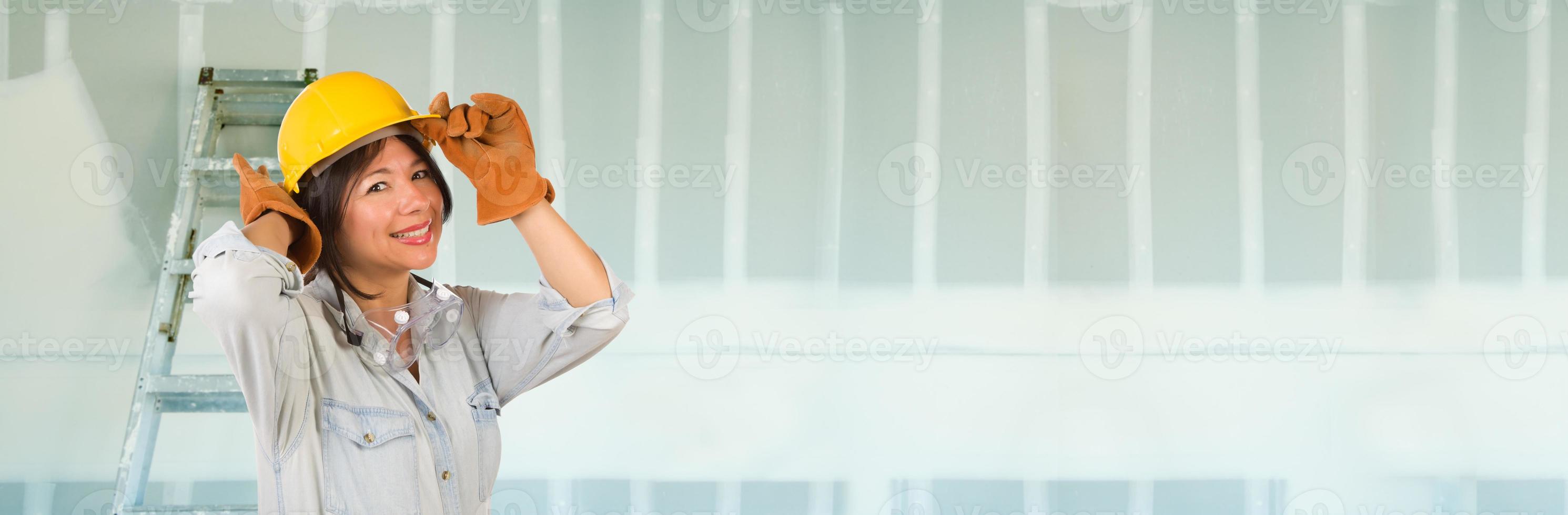 Hispanic Female Contractor Wearing Hard Hat Against Drywall Banner Background with Ladder photo