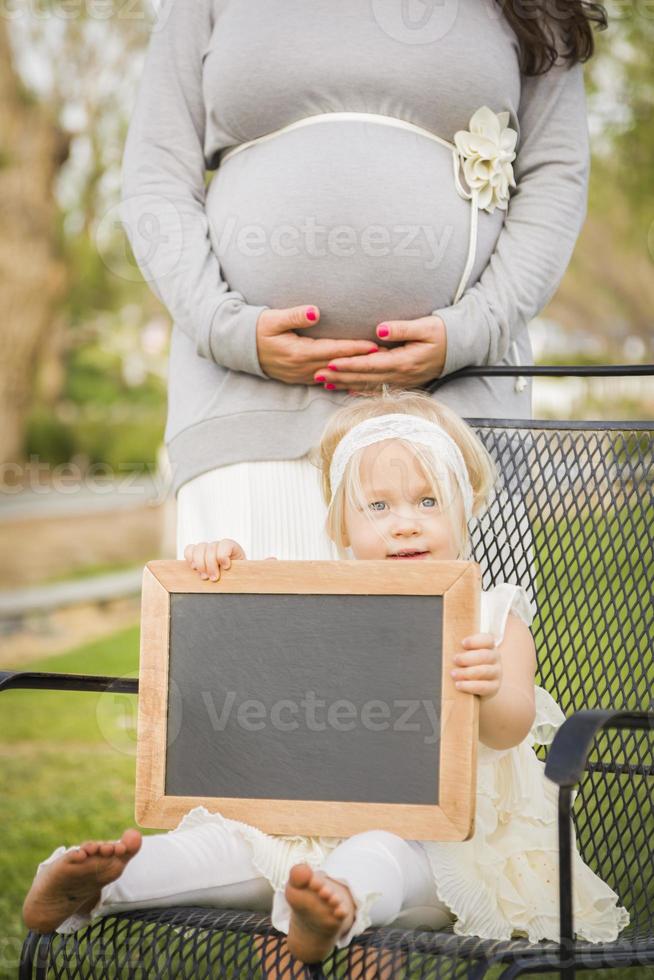 mamá embarazada detrás de una niña en una silla sosteniendo una pizarra en blanco foto