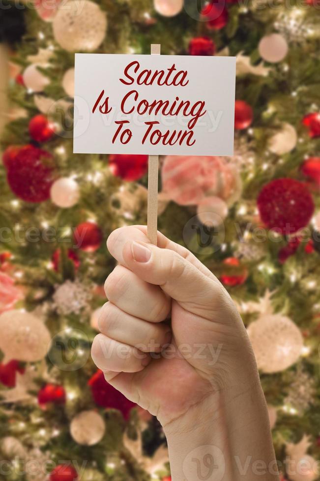 la mano que sostiene a santa viene a la tarjeta de la ciudad frente al árbol de navidad decorado. foto