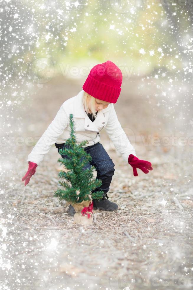 chica con mitones rojos y gorra cerca de un pequeño árbol de navidad con efecto de nieve foto