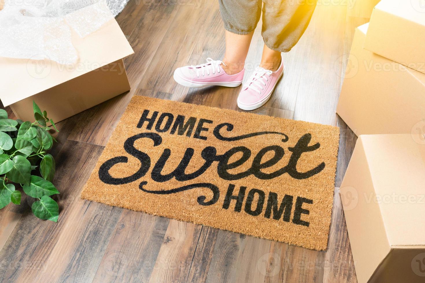 Woman in Pink Shoes and Sweats Standing Near Home Sweet Home Welcome Mat, Boxes and Plant photo