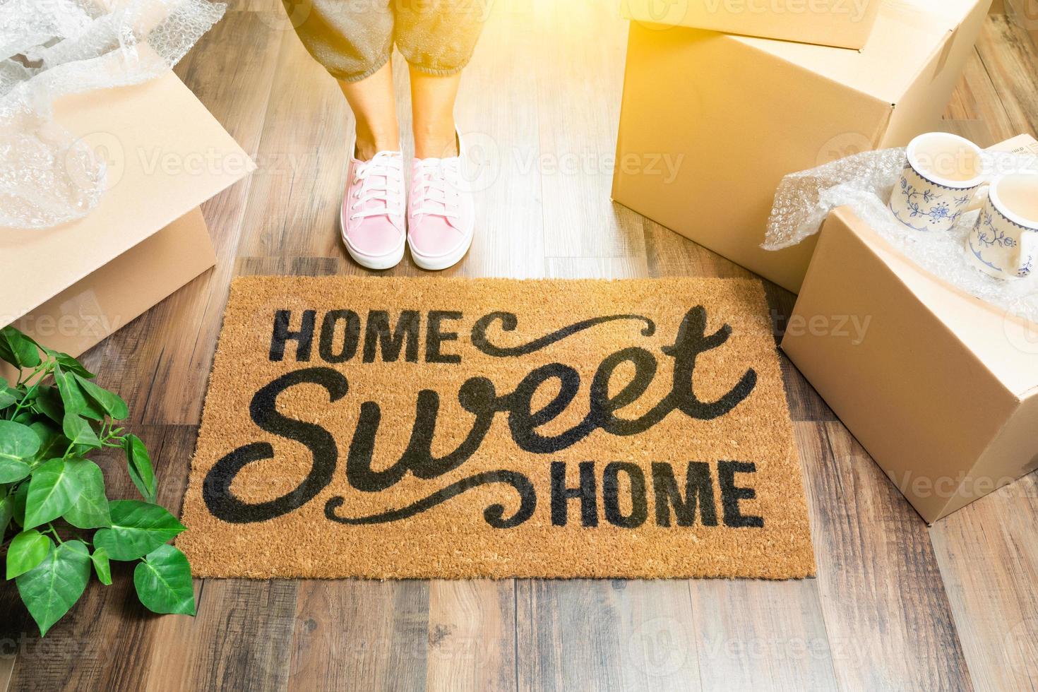 Woman in Pink Shoes and Sweats Standing Near Home Sweet Home Welcome Mat, Boxes and Plant photo