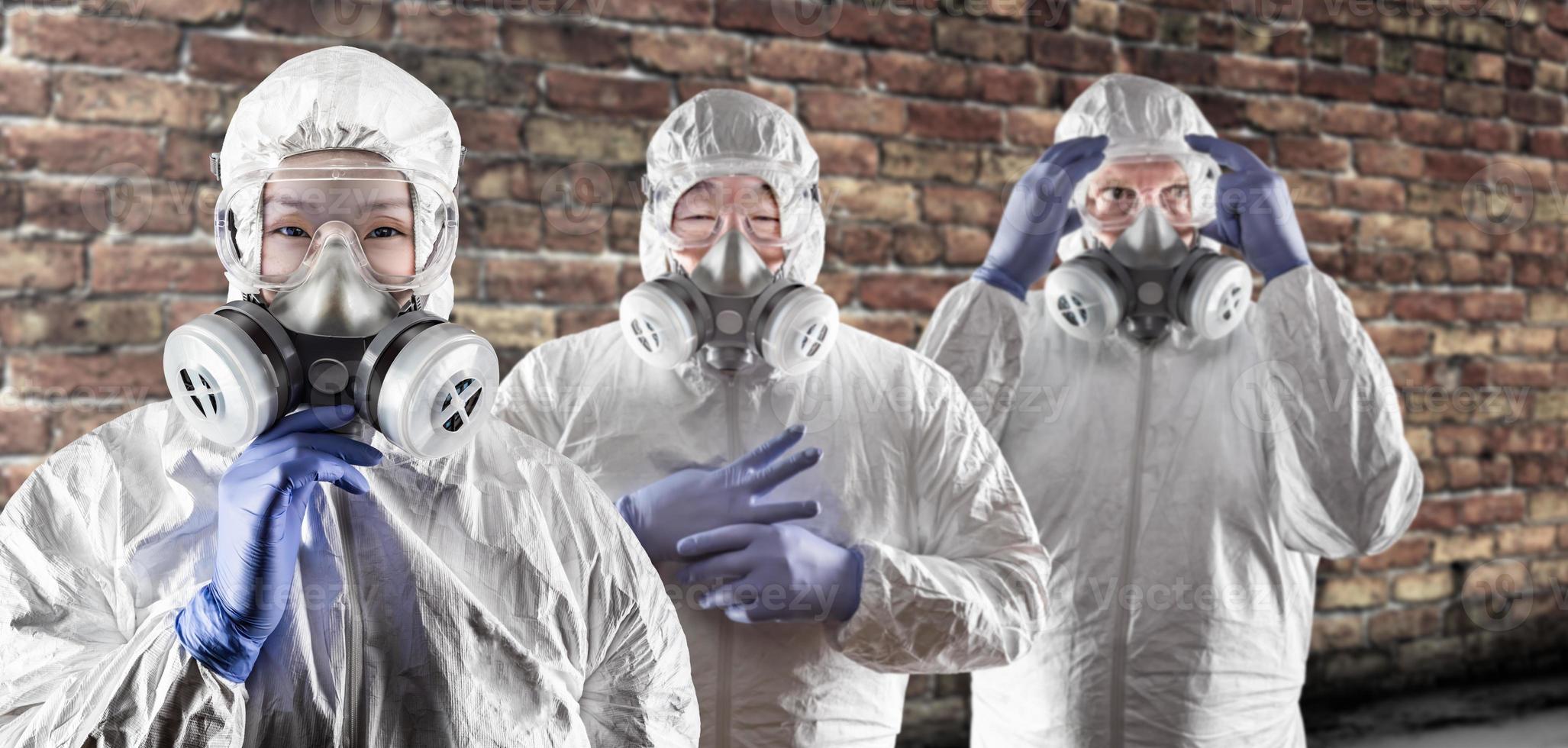Chinese and Caucasian Woman and Men In Gas Masks, Goggles and Hazmat Suites Against Brick Background photo