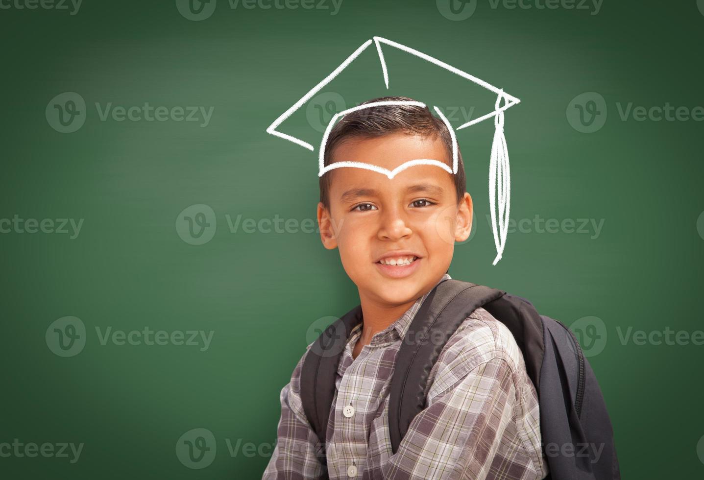 Young Hispanic Student Boy Wearing Backpack Front Of Blackboard with Graduation Cap Drawn In Chalk Over Head photo