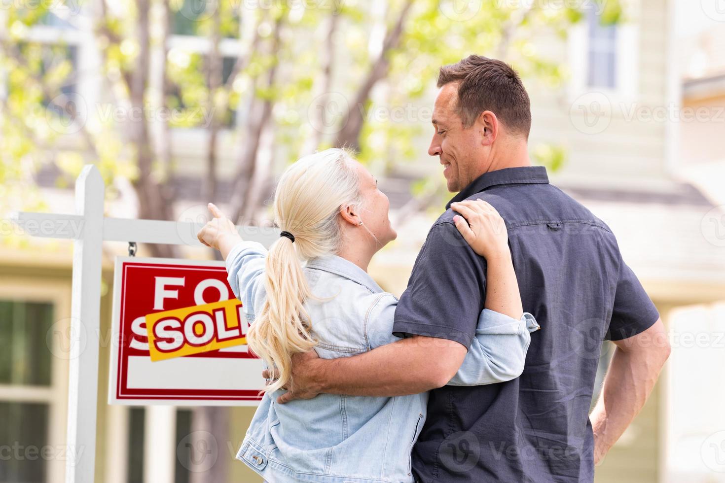 Caucasian Couple Facing and Pointing to Front of Sold Real Estate Sign and House. photo