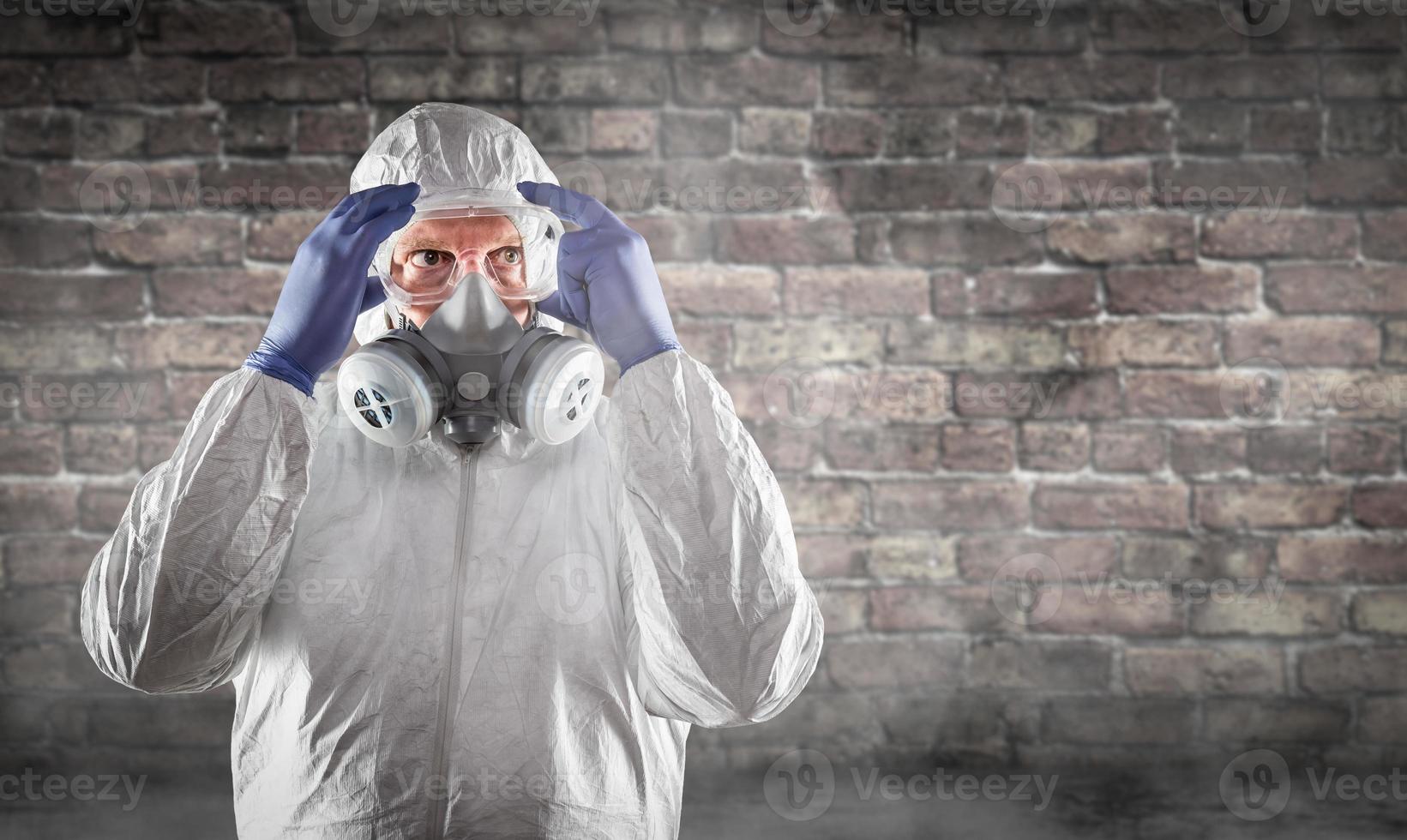 Man Wearing Hazmat Suit, Protective Gas Mask and Goggles Against Brick Wall photo