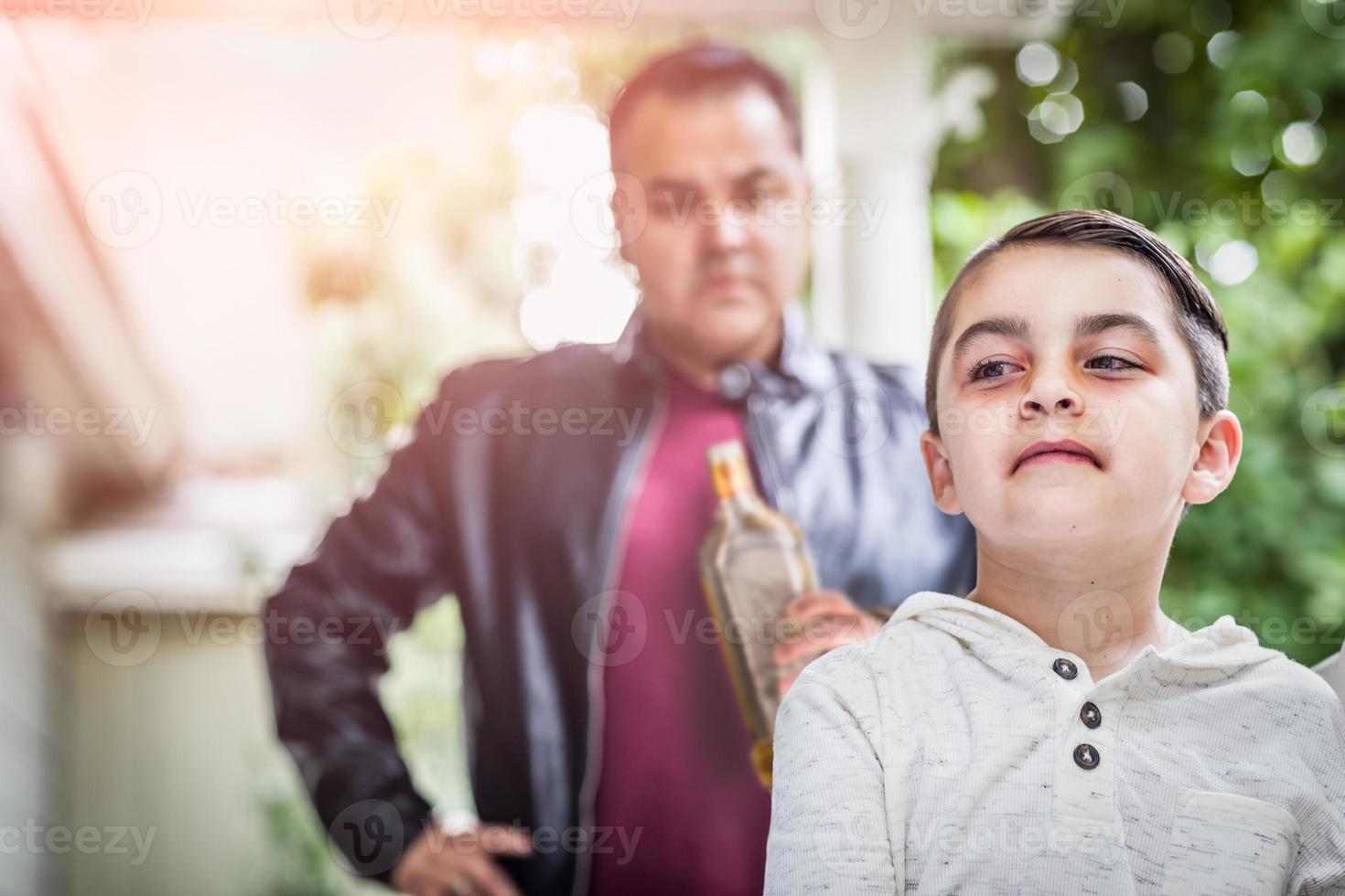 Afraid and Bruised Mixed Race Boy In Front of Angry Man Holding Bottle of Alcohol photo