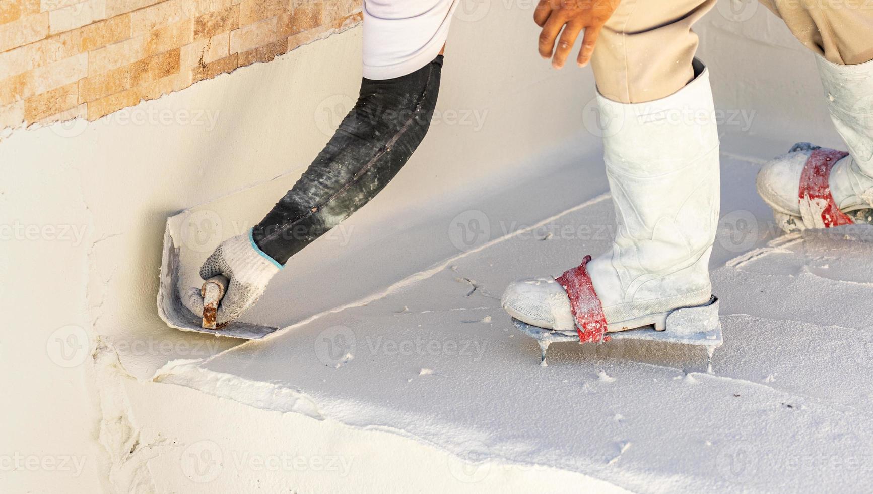 trabajador que usa zapatos con púas alisando el yeso húmedo de la piscina con una paleta foto