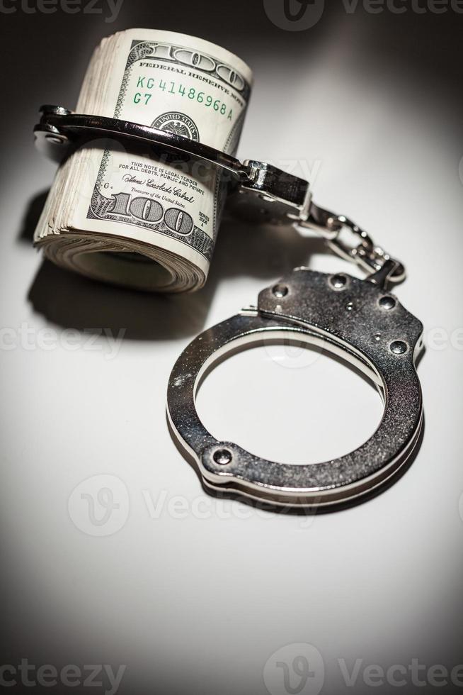 Handcuffs Locked on Roll of One Hundred Dollar Bills Under Spotlight photo