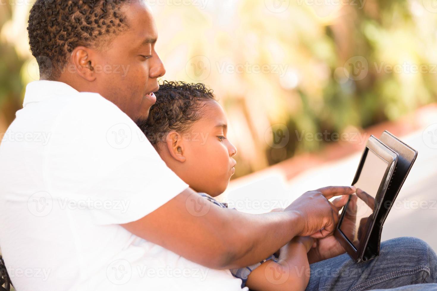 African American Father and Mixed Race Son Using Computer Tablet on Bench in Park photo