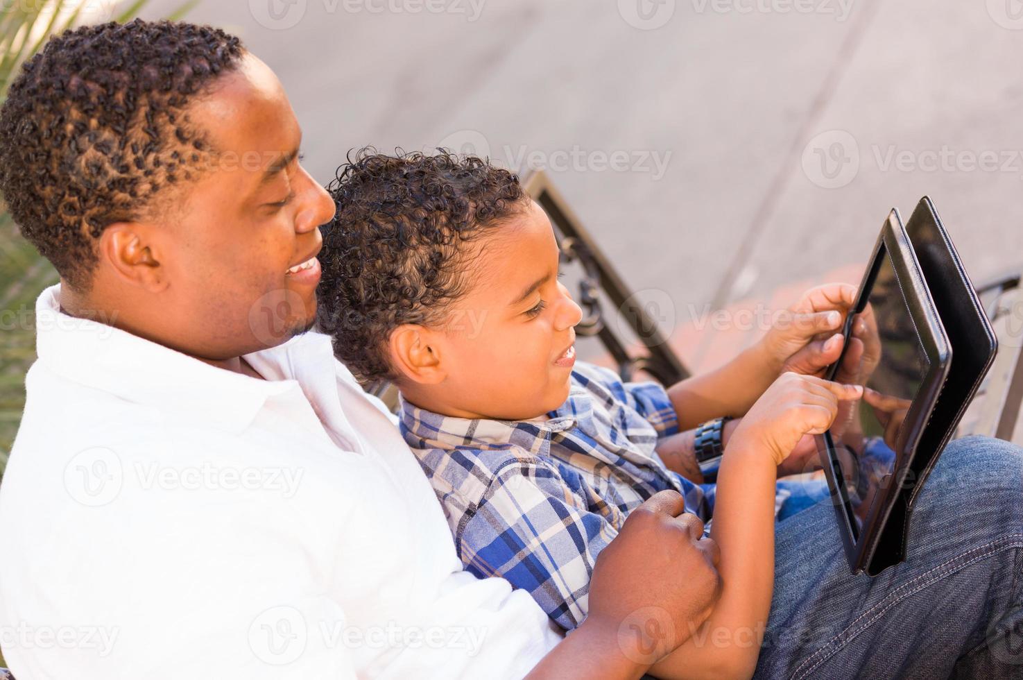 African American Father and Mixed Race Son Using Computer Tablet on Bench in Park photo